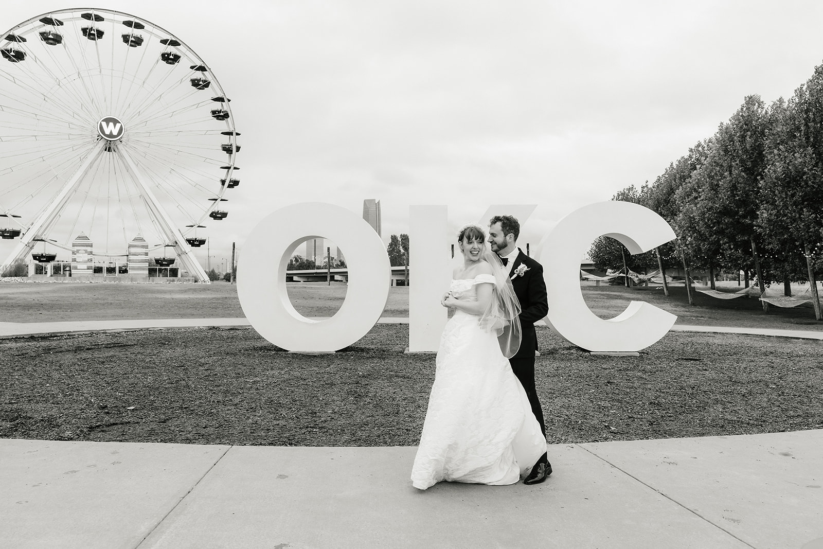 A bride and groom stand in front of large "OKC" letters and a Ferris wheel, holding hands outdoors on a cloudy day.