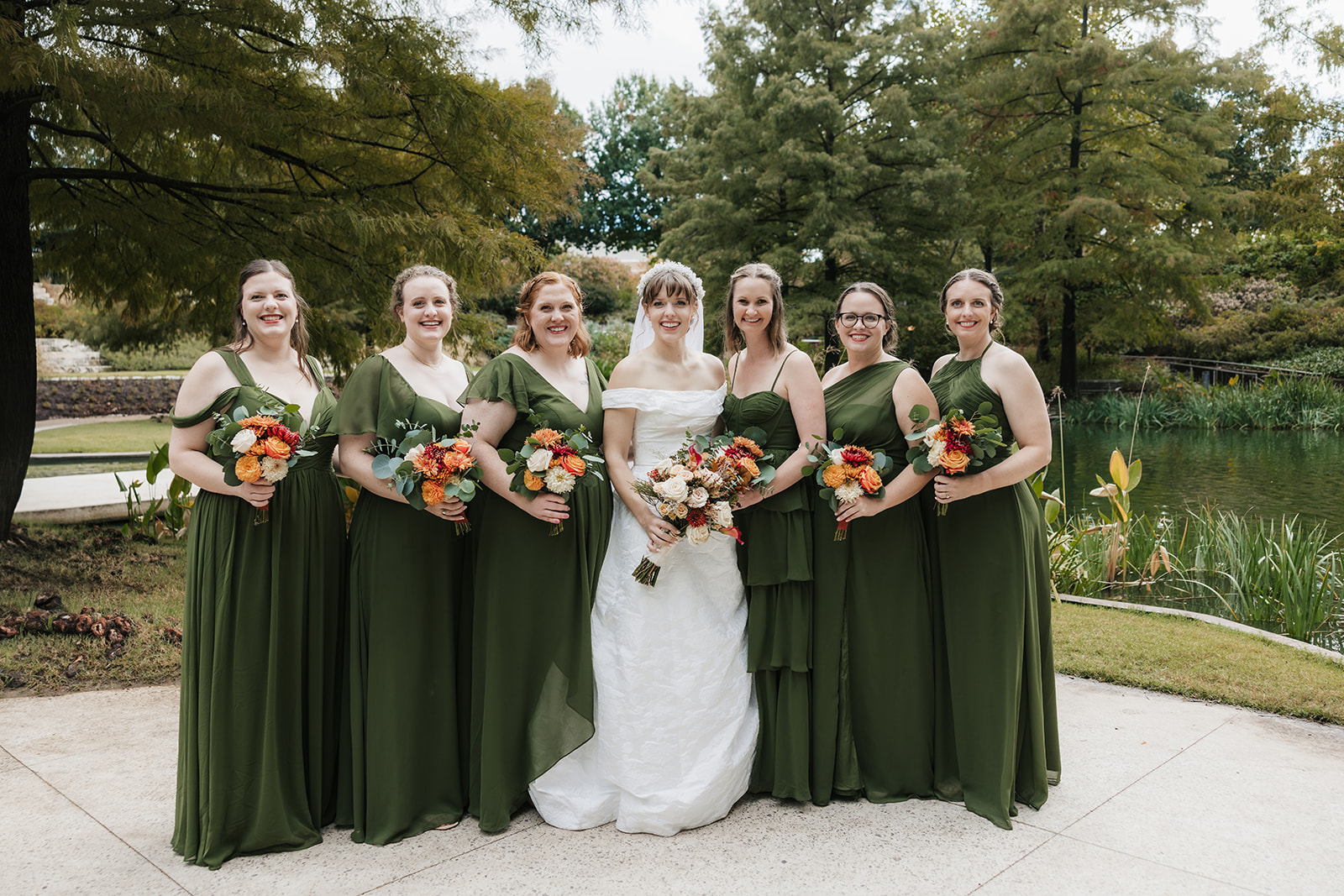 Seven women stand outdoors, with one in a white wedding dress and six in matching green dresses, all holding bouquets of flowers. Trees and a pond are visible in the background captured by a catholic wedding photographer