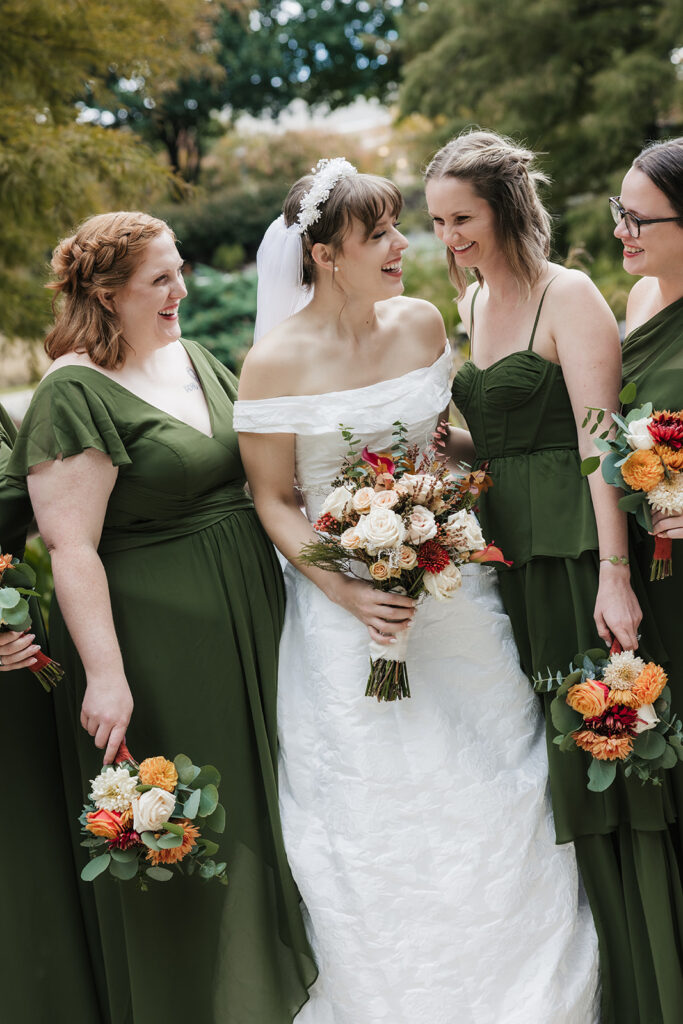Seven women stand outdoors, with one in a white wedding dress and six in matching green dresses, all holding bouquets of flowers. Trees and a pond are visible in the background captured by a catholic wedding photographer