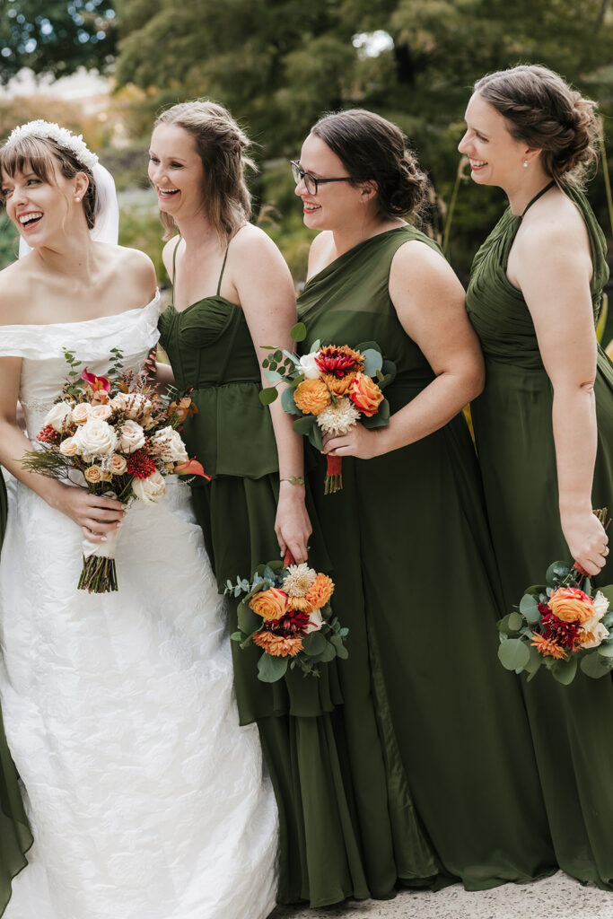 Seven women stand outdoors, with one in a white wedding dress and six in matching green dresses, all holding bouquets of flowers. Trees and a pond are visible in the background captured by a catholic wedding photographer