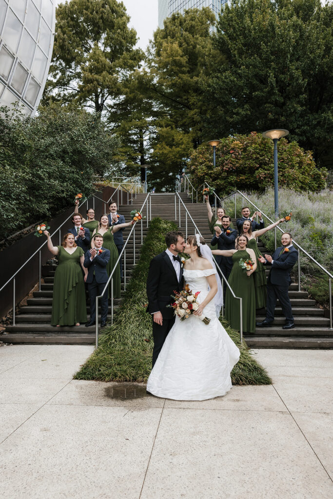A bride and groom kiss at the base of outdoor stairs while their wedding party, dressed in green and black, stands on the steps holding bouquets.