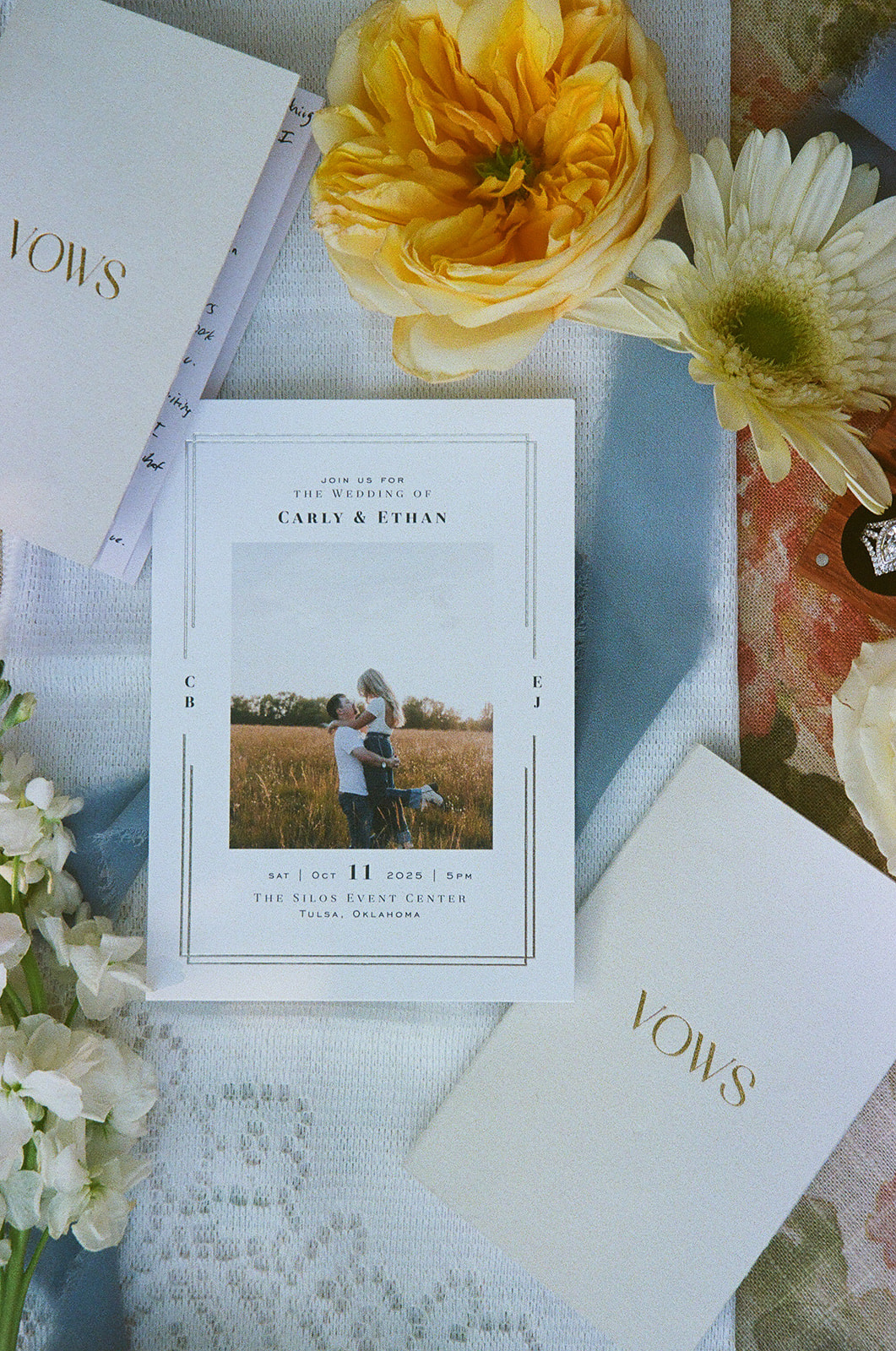 A wedding invitation with a photo of a couple is surrounded by flowers and two white booklets labeled “Vows” in gold lettering for a wedding at The Silo Event Center in Tulsa
