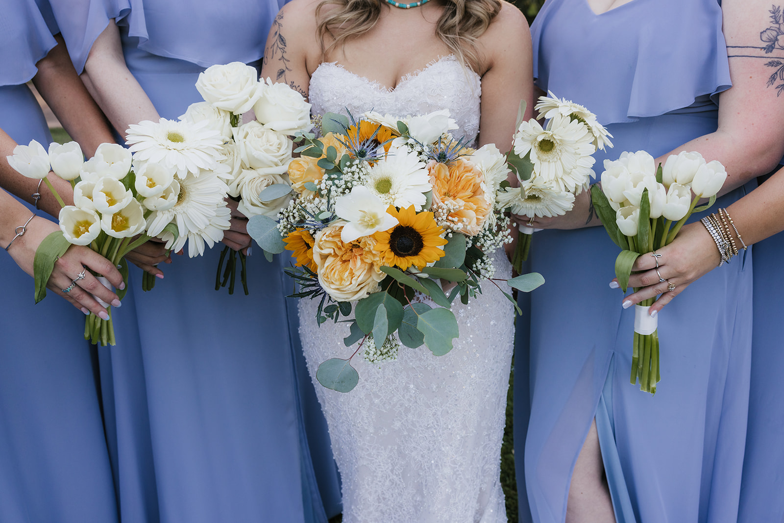 A bride in a white gown stands outdoors with four bridesmaids in matching light blue dresses, all holding bouquets and smiling at the Silo Event Center in Tulsa