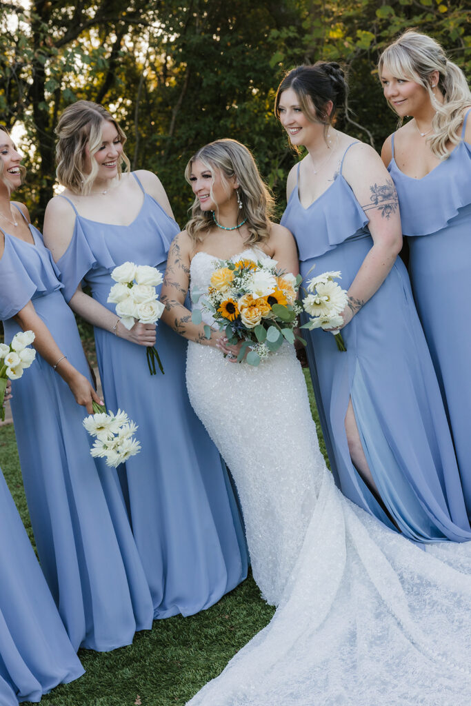 A bride in a white gown stands outdoors with four bridesmaids in matching light blue dresses, all holding bouquets and smiling at the Silo Event Center in Tulsa