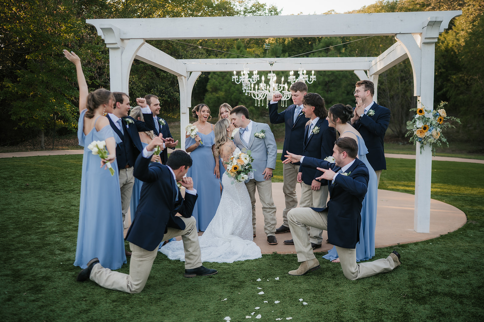 A wedding party poses outdoors under a white pergola; the bridesmaids wear light blue dresses, and the groomsmen wear dark jackets and khaki pants.