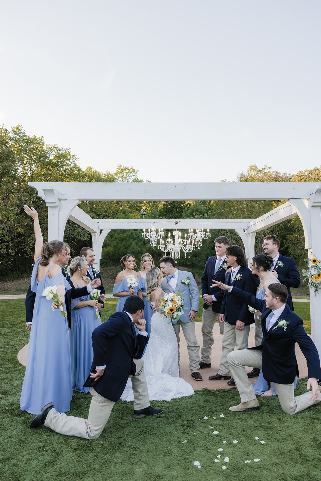 A bride in a white gown stands outdoors with four bridesmaids in matching light blue dresses, all holding bouquets and smiling at the Silo Event Center in Tulsa