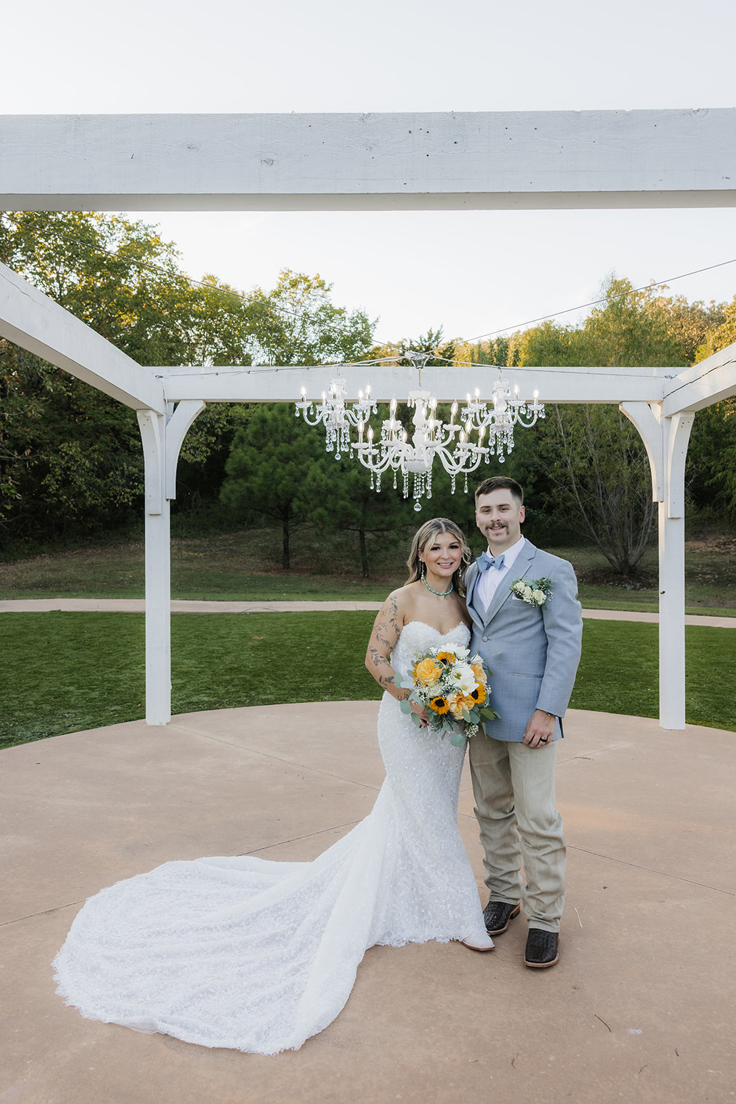 A bride and groom kiss under a white wooden pergola with a chandelier, surrounded by greenery on a circular platform at The Silo Event Center in Tulsa