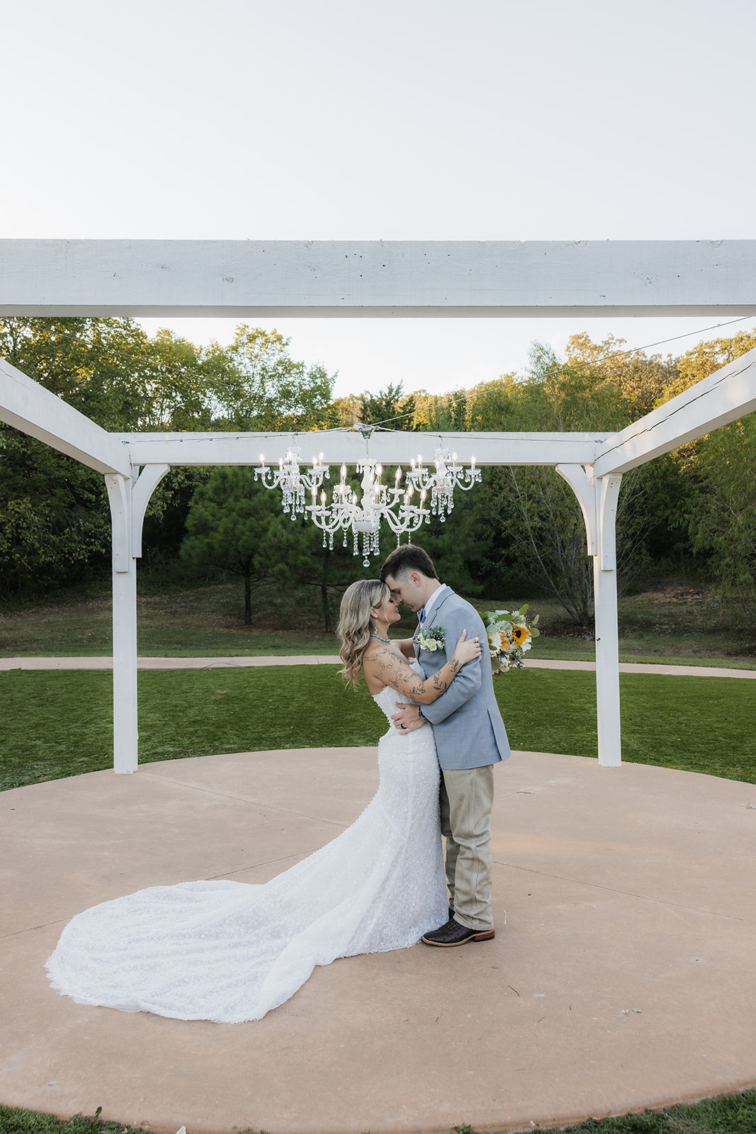 A bride and groom kiss under a white wooden pergola with a chandelier, surrounded by greenery on a circular platform at The Silo Event Center in Tulsa
