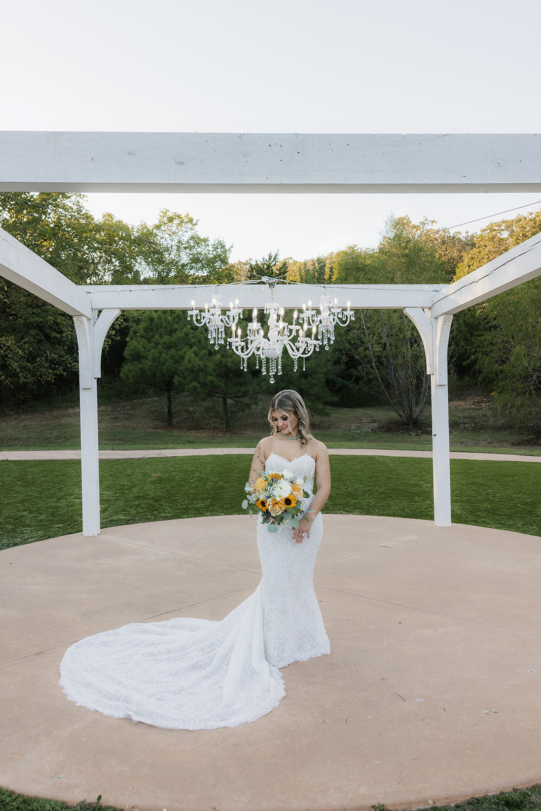 A bride in a strapless white dress holding a sunflower bouquet sits on a white chair outdoors, surrounded by empty chairs.