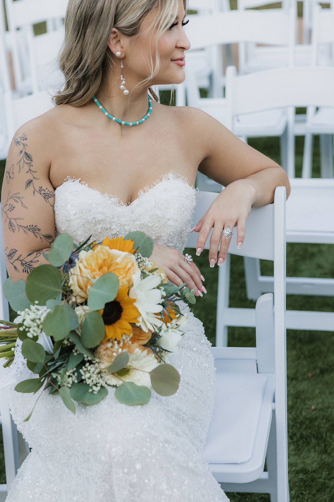 A bride in a strapless white dress holding a sunflower bouquet sits on a white chair outdoors, surrounded by empty chairs.