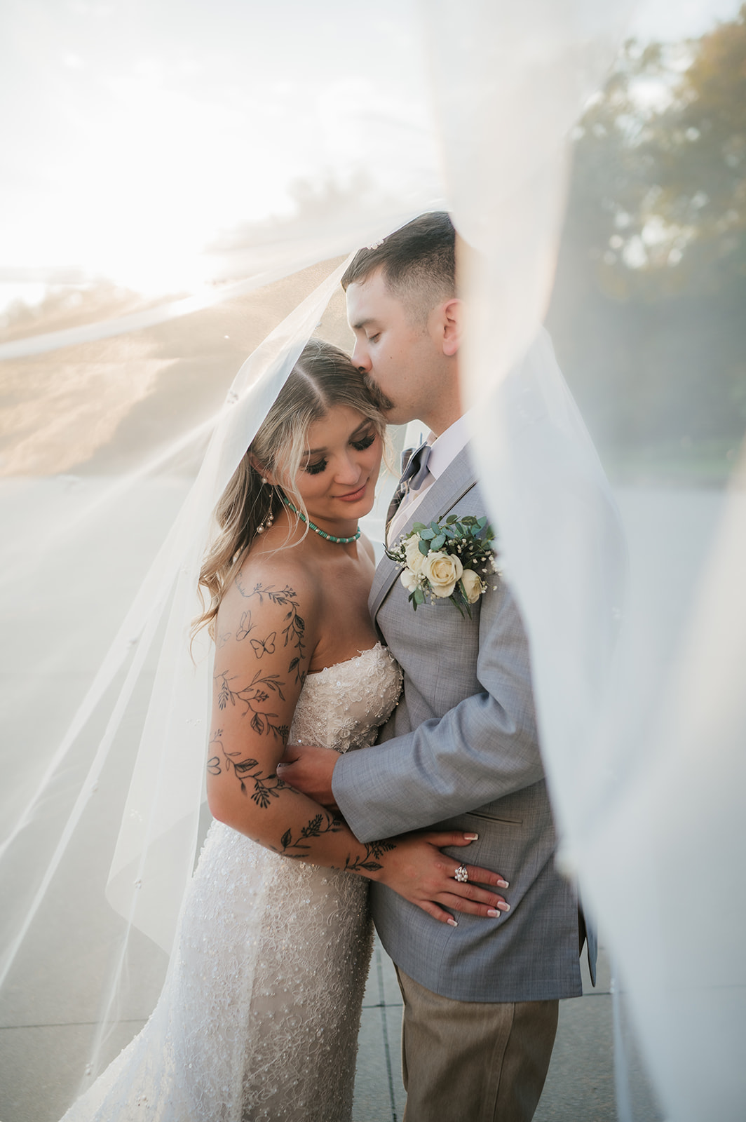 A bride and groom stand close together under a flowing veil outdoors; the bride wears a white dress and the groom a light gray suit with a floral boutonniere at aWedding at The Silo Event Center in Tulsa