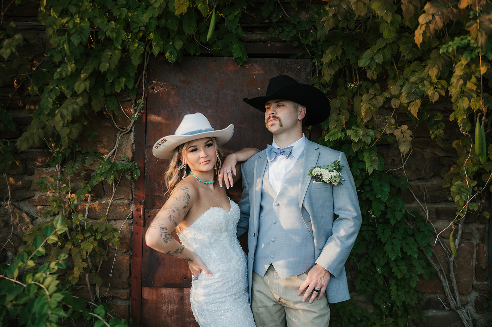 A bride and groom in western attire pose together in front of a rustic door covered with green vines.