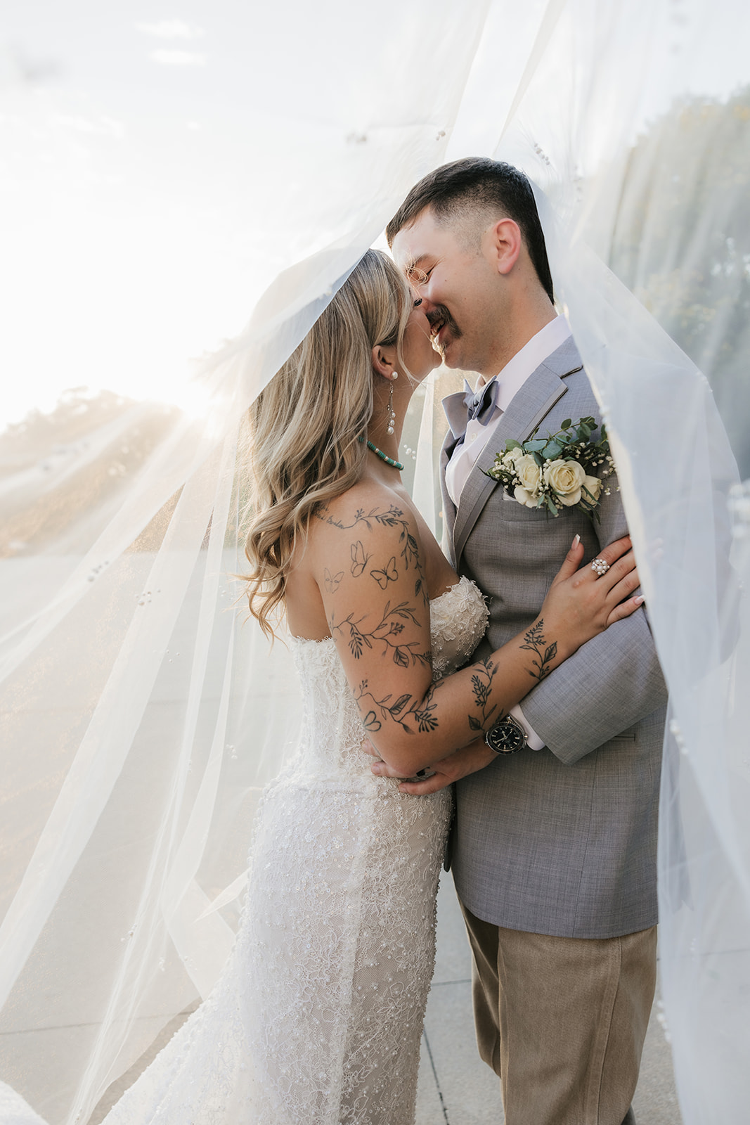 A bride and groom stand close together under a flowing veil outdoors; the bride wears a white dress and the groom a light gray suit with a floral boutonniere at aWedding at The Silo Event Center in Tulsa