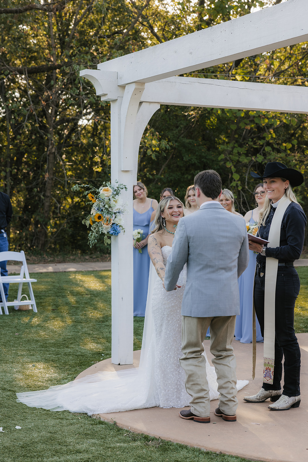 A bride and groom kiss under a white pergola during an outdoor wedding ceremony, with guests and an officiant present at The Silo Event Center in Tulsa