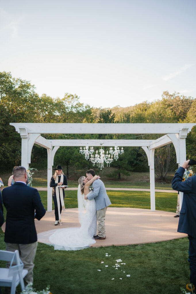 A bride and groom kiss under a white pergola during an outdoor wedding ceremony, with guests and an officiant present at The Silo Event Center in Tulsa