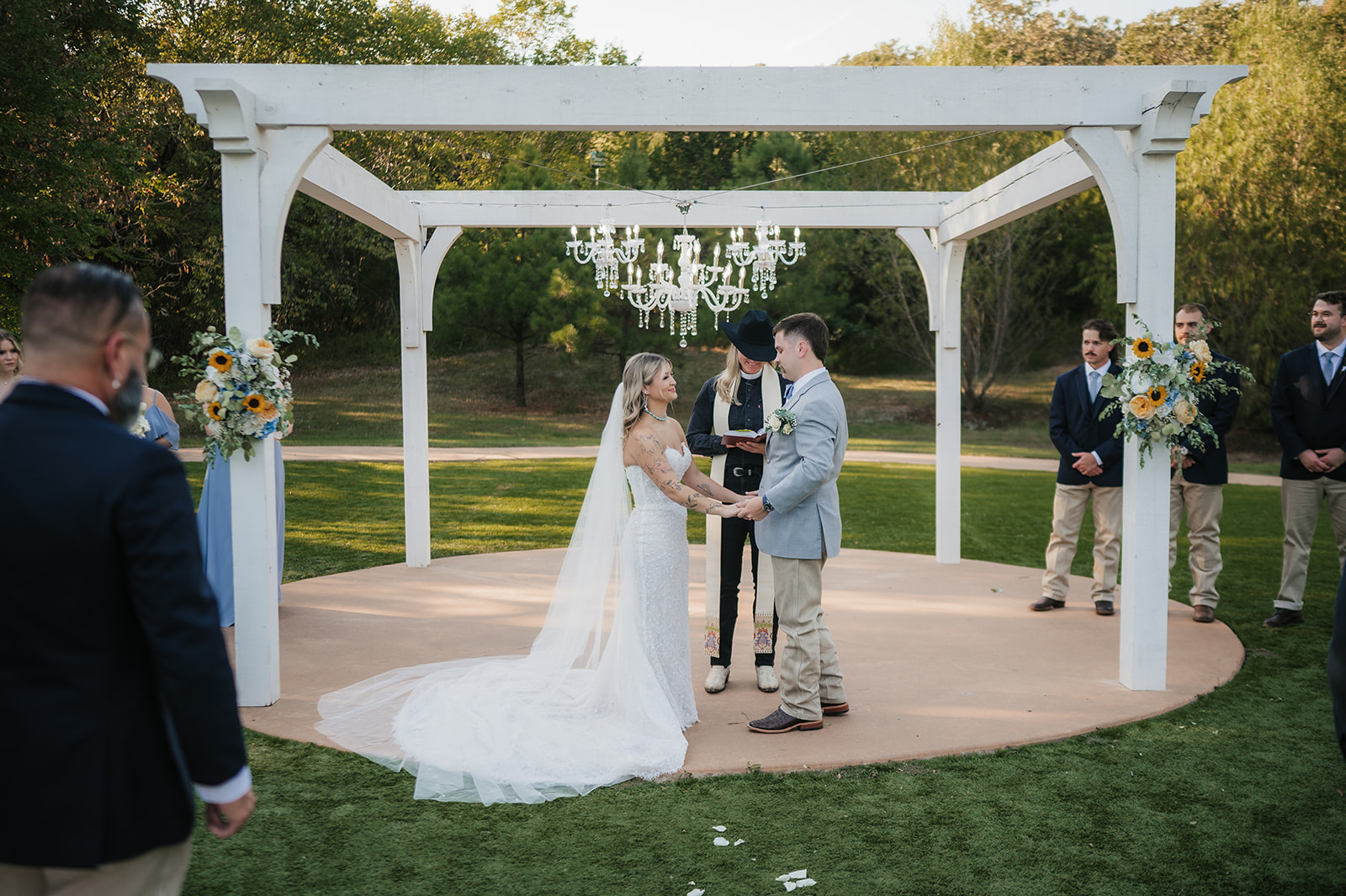 A bride and groom kiss under a white pergola during an outdoor wedding ceremony, with guests and an officiant present at The Silo Event Center in Tulsa