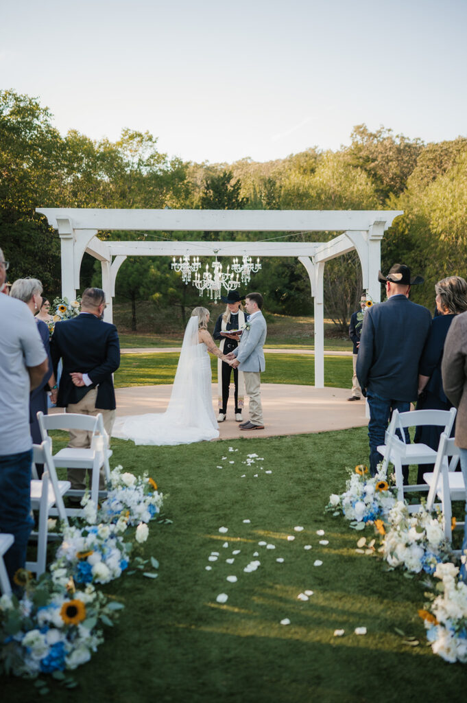 A bride and groom kiss under a white pergola during an outdoor wedding ceremony, with guests and an officiant present at The Silo Event Center in Tulsa