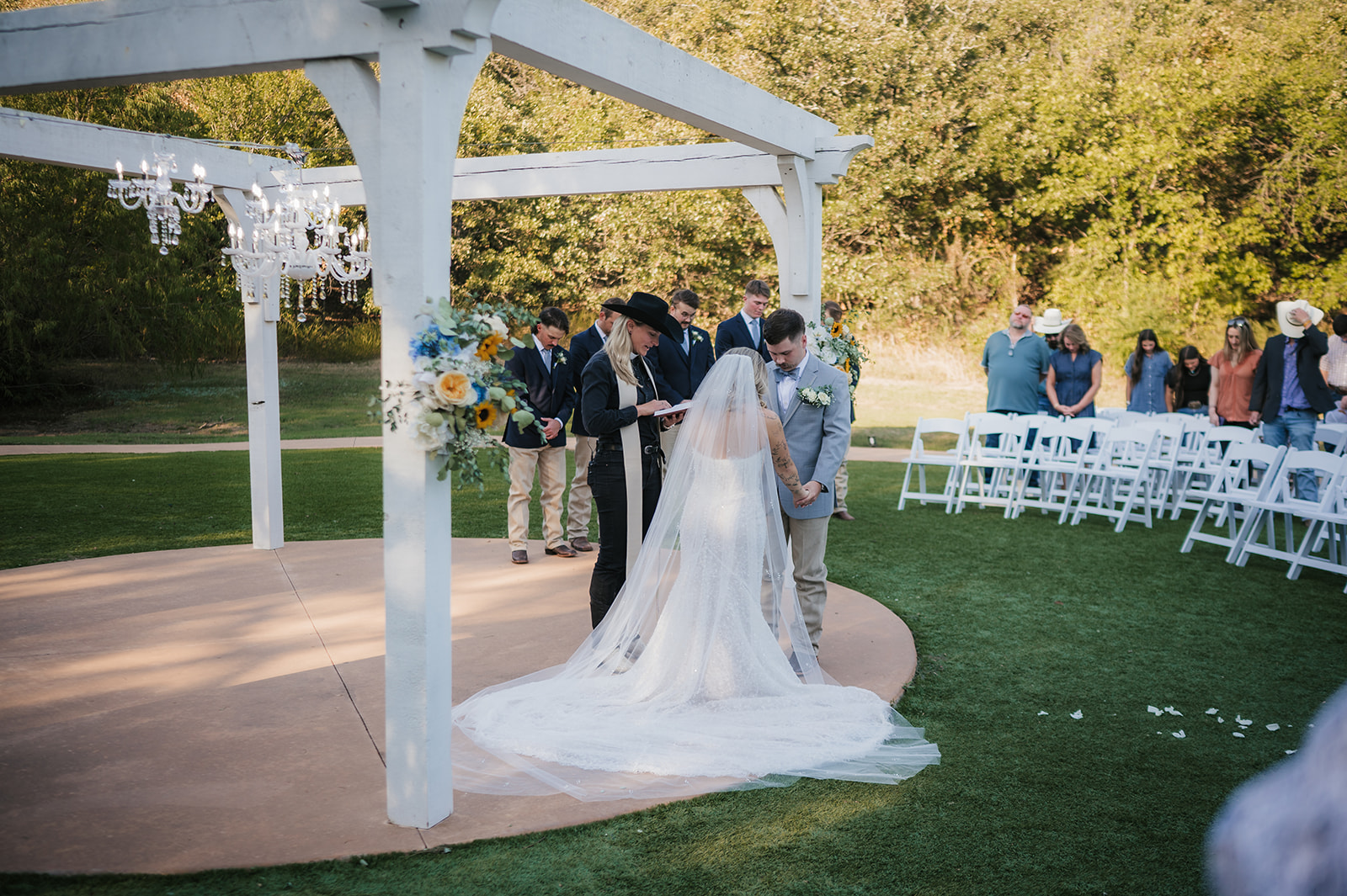 A bride and groom kiss under a white pergola during an outdoor wedding ceremony, with guests and an officiant present at The Silo Event Center in Tulsa