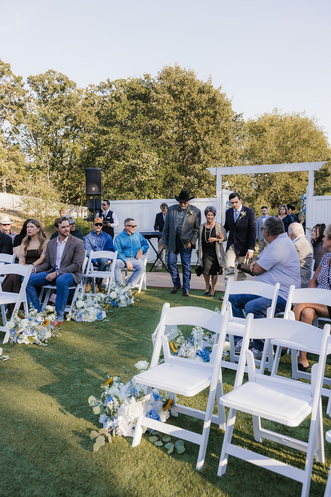 A bride and groom kiss under a white pergola during an outdoor wedding ceremony, with guests and an officiant present at The Silo Event Center in Tulsa