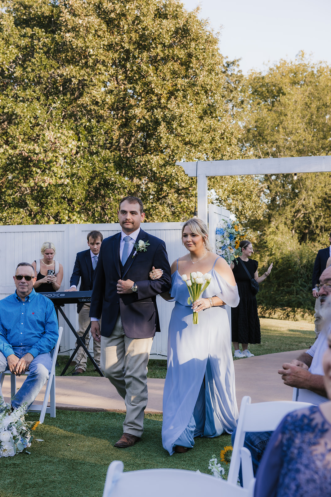 A bride and groom kiss under a white pergola during an outdoor wedding ceremony, with guests and an officiant present at The Silo Event Center in Tulsa