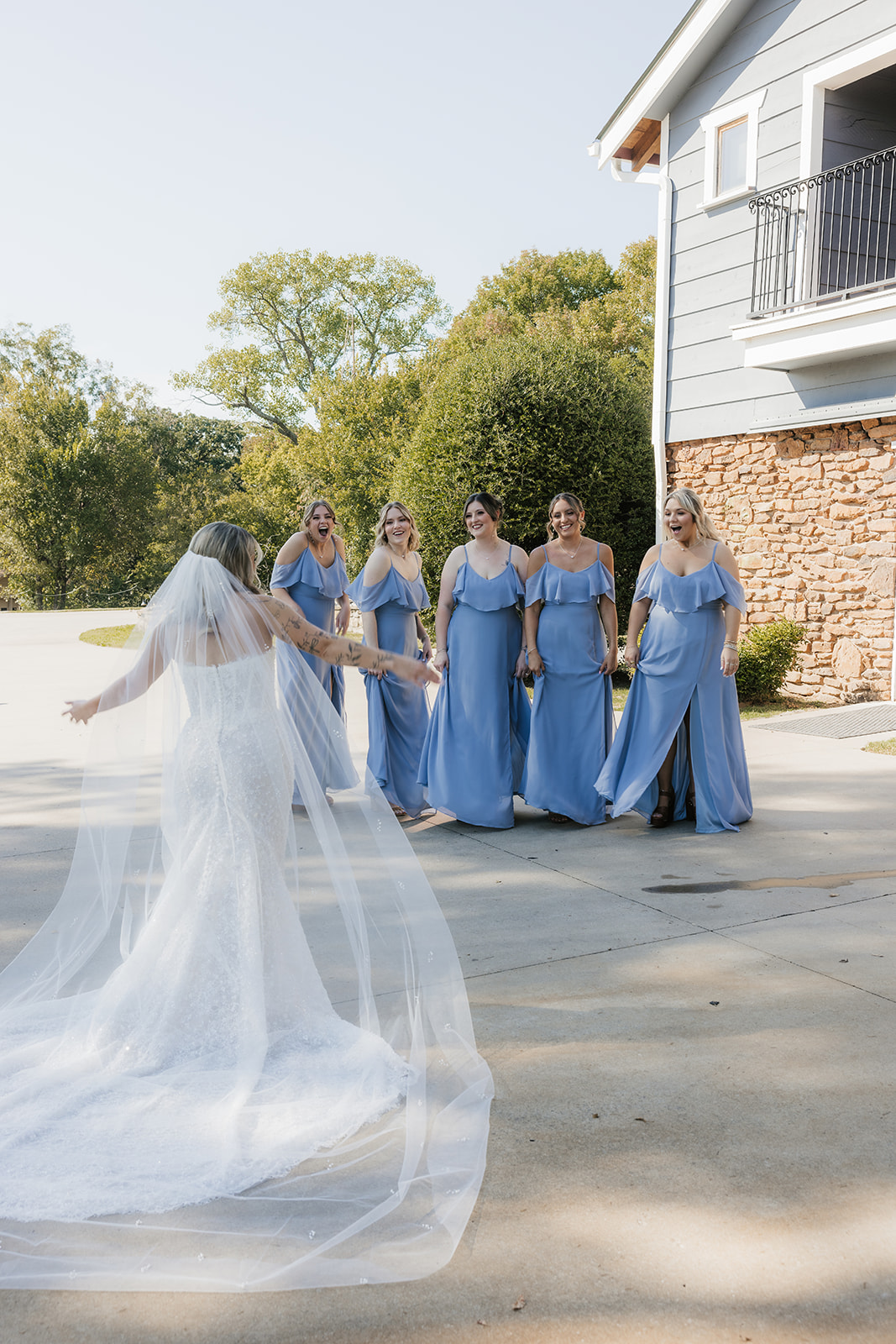 A bride in a white gown and veil stands facing five bridesmaids in matching blue dresses outside near a house at The Silo Event Center in Tulsa