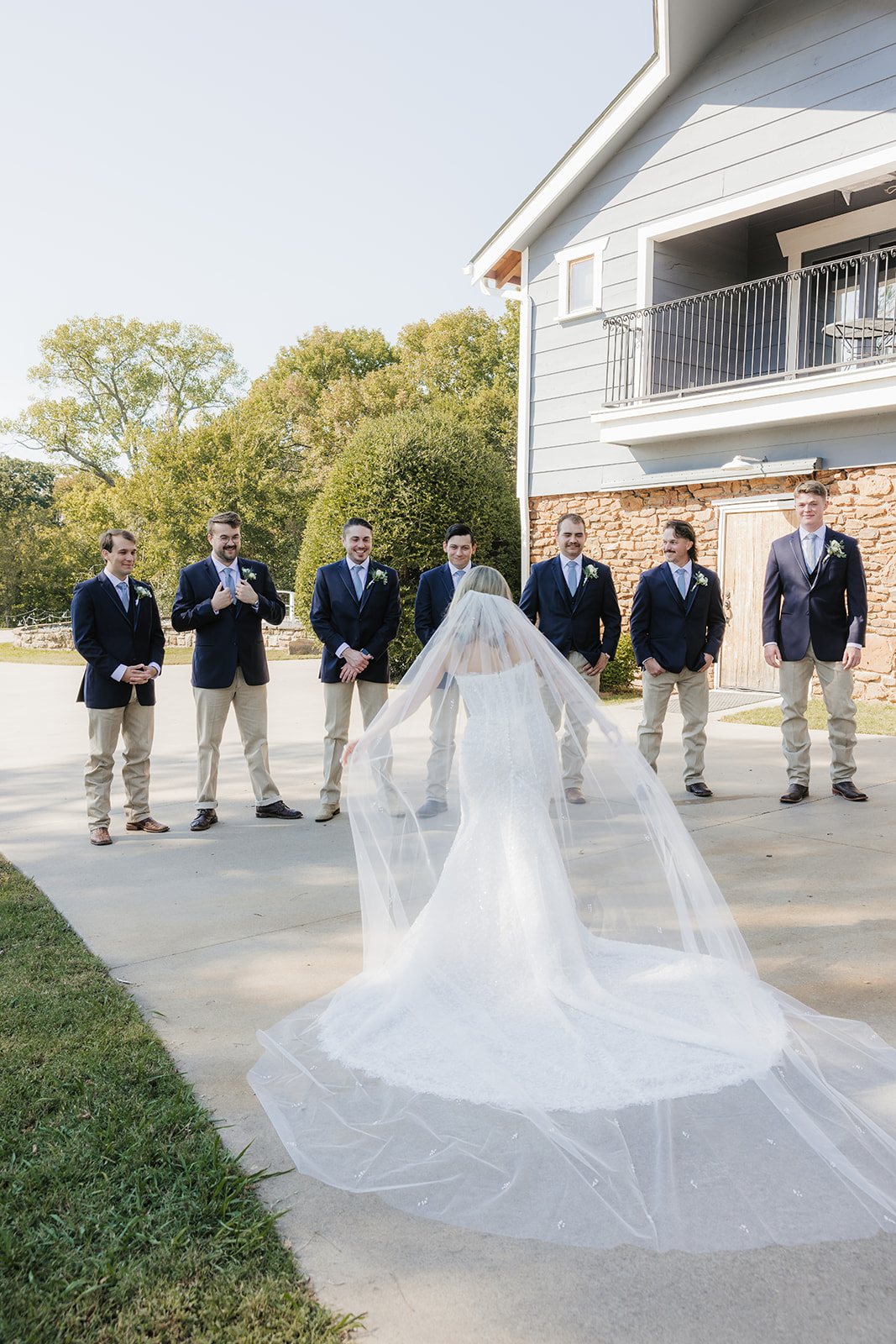 A bride in a white gown and veil stands facing five bridesmaids in matching blue dresses outside near a house at The Silo Event Center in Tulsa