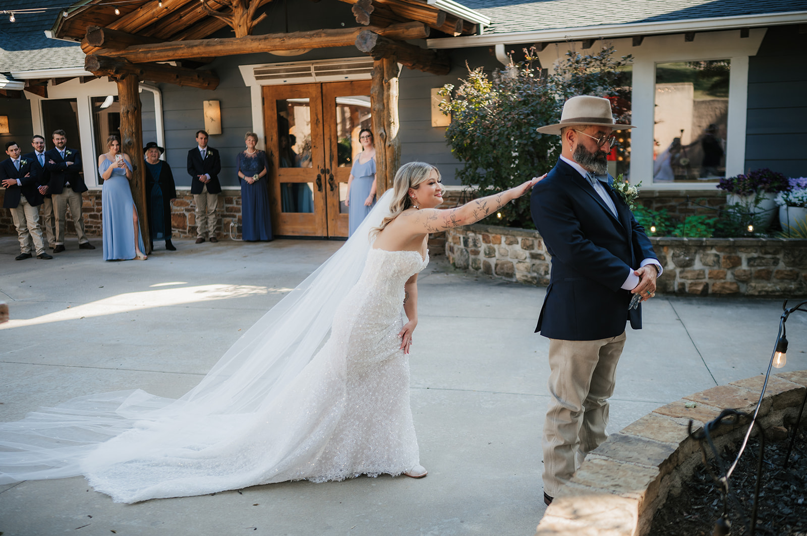 A bride in a white gown and veil stands facing five bridesmaids in matching blue dresses outside near a house at The Silo Event Center in Tulsa