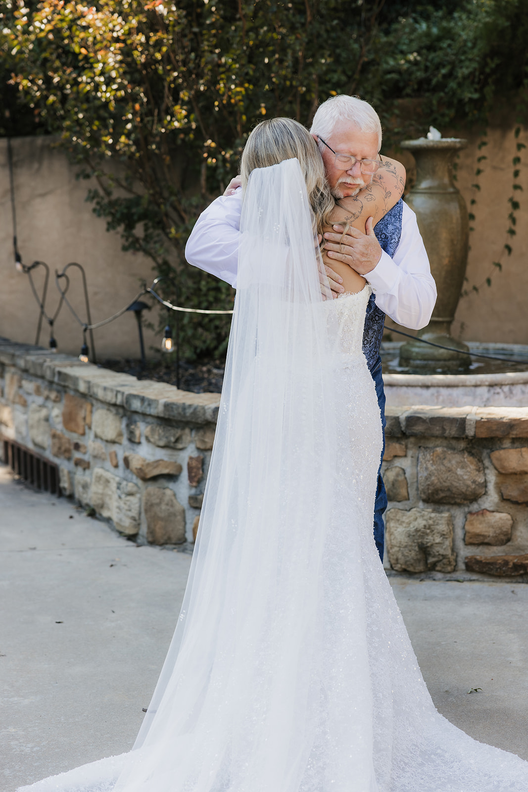 A bride in a white gown and veil stands facing five bridesmaids in matching blue dresses outside near a house at The Silo Event Center in Tulsa