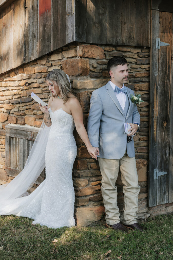 A bride and groom stand back-to-back outside a rustic building, holding hands. The bride wears a white gown and veil; the groom holds a piece of paper and wears a light-colored suit. for a wedding at The Silo Event Center in Tulsa