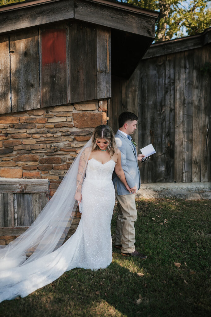 A bride and groom stand back-to-back outside a rustic building, holding hands. The bride wears a white gown and veil; the groom holds a piece of paper and wears a light-colored suit. for a wedding at The Silo Event Center in Tulsa