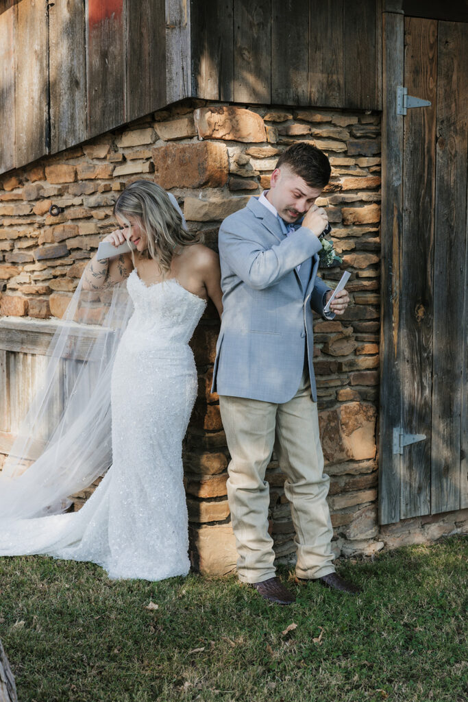 A bride and groom stand back-to-back outside a rustic building, holding hands. The bride wears a white gown and veil; the groom holds a piece of paper and wears a light-colored suit. for a wedding at The Silo Event Center in Tulsa