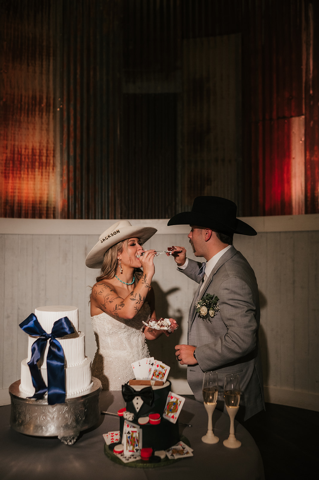 A bride and groom wearing cowboy hats feed each other cake beside a decorated wedding cake and glasses of champagne