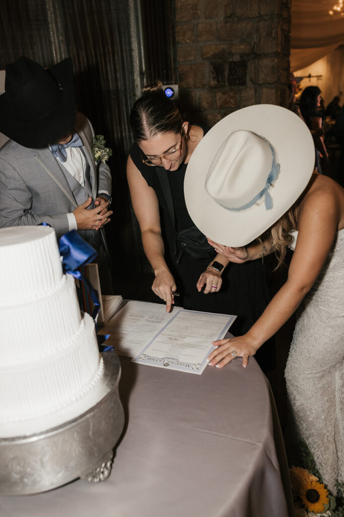 Three people stand around a table, with two signing a document next to a white tiered cake. One person wears a wedding dress and hat.