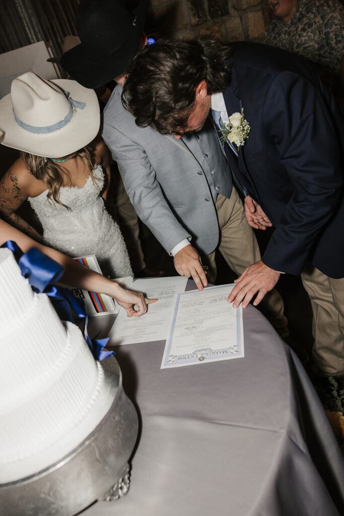 Three people stand around a table, with two signing a document next to a white tiered cake. One person wears a wedding dress and hat.
