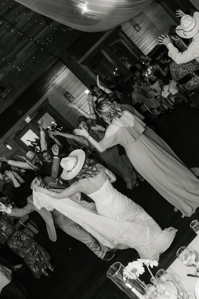 A bride in a wedding dress dances among guests, some wearing hats, in a lively indoor celebration.