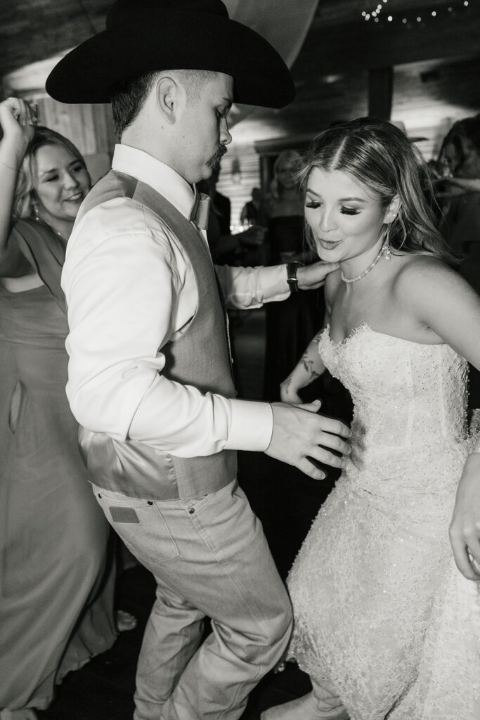 A bride in a wedding dress dances among guests, some wearing hats, in a lively indoor celebration.