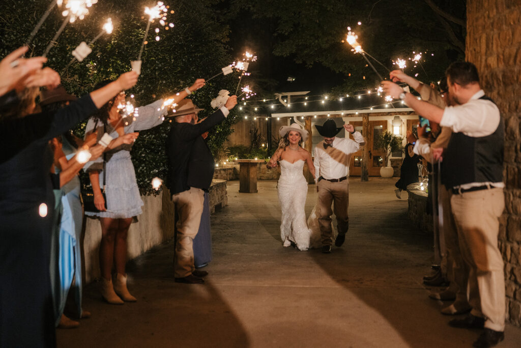 A bride and groom walk down a path at night, surrounded by guests holding sparklers and celebrating.