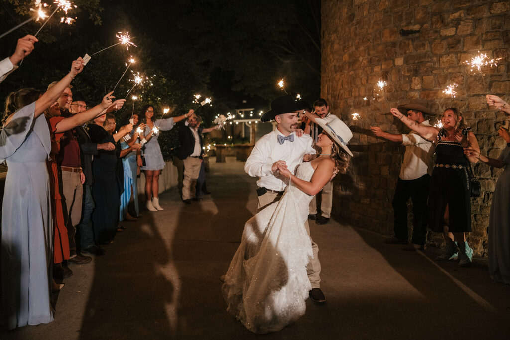 A bride and groom walk down a path at night, surrounded by guests holding sparklers and celebrating.