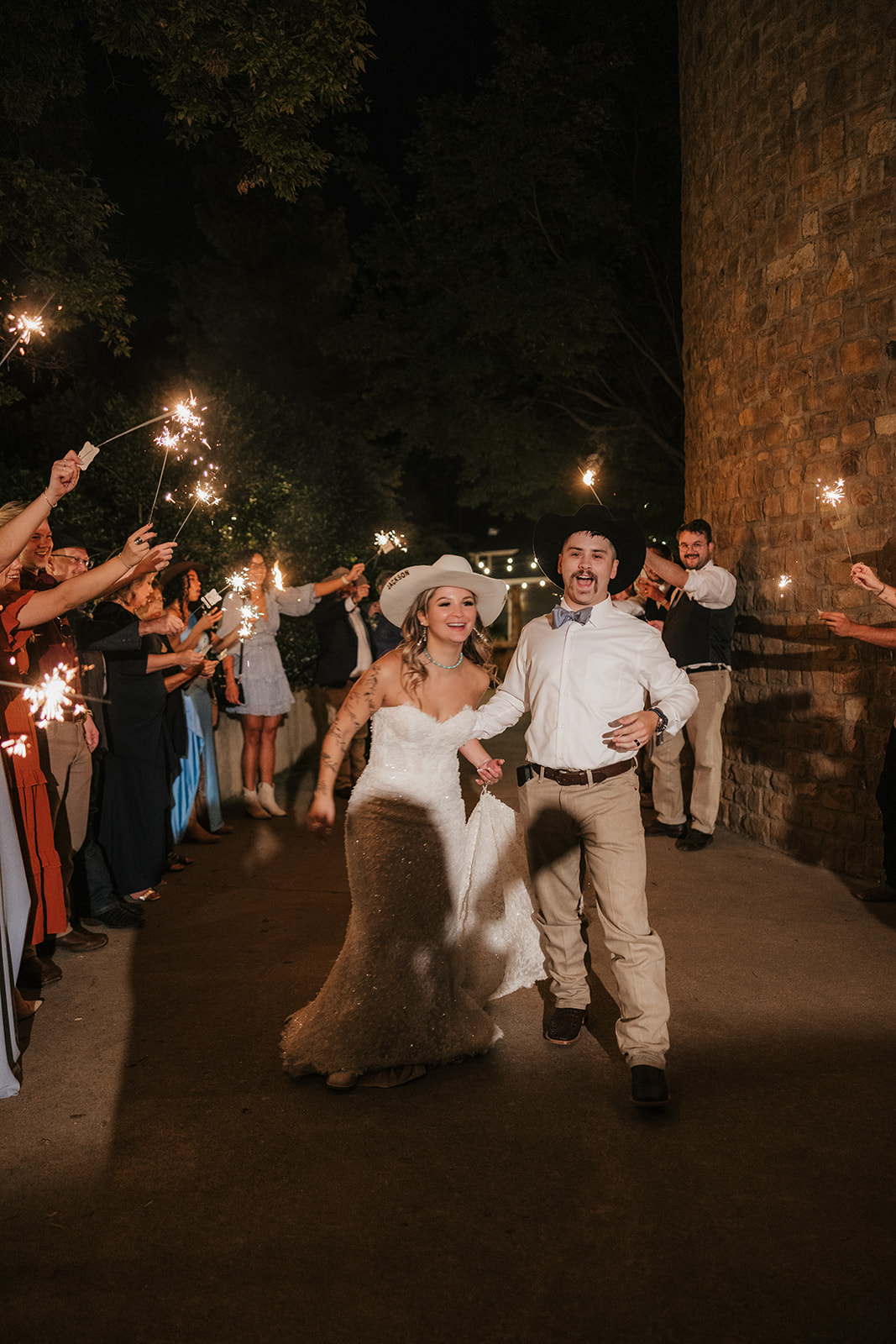 A bride and groom walk down a path at night, surrounded by guests holding sparklers and celebrating.