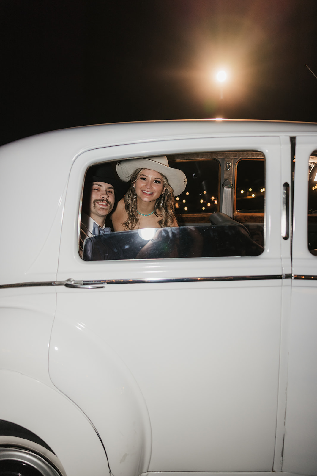 Two people smiling and looking out the back window of a white vintage car at night, with streetlights visible in the background.