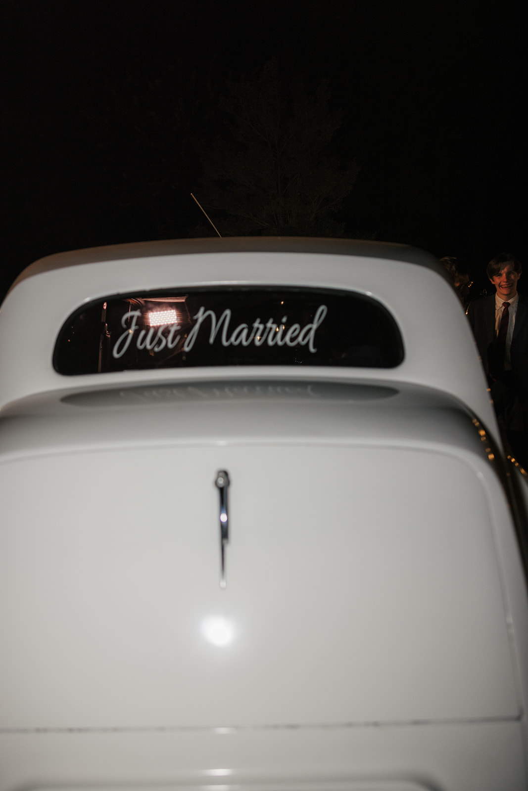 Two people smiling and looking out the back window of a white vintage car at night, with streetlights visible in the background.