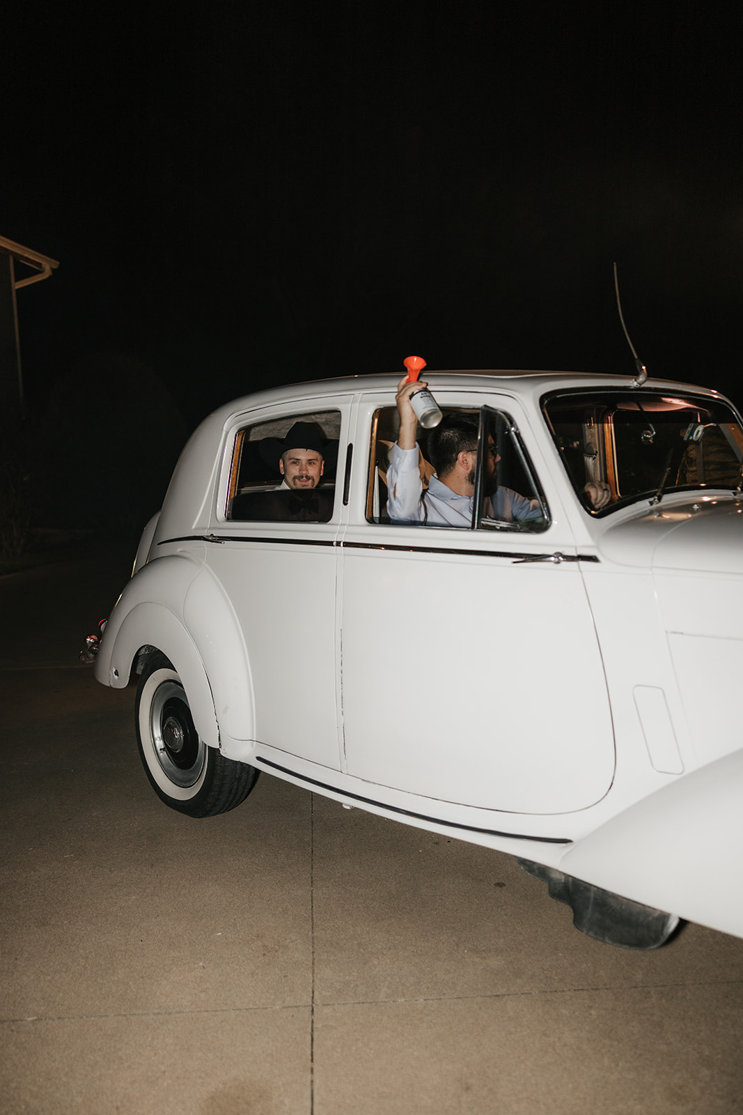 Two people smiling and looking out the back window of a white vintage car at night, with streetlights visible in the background.