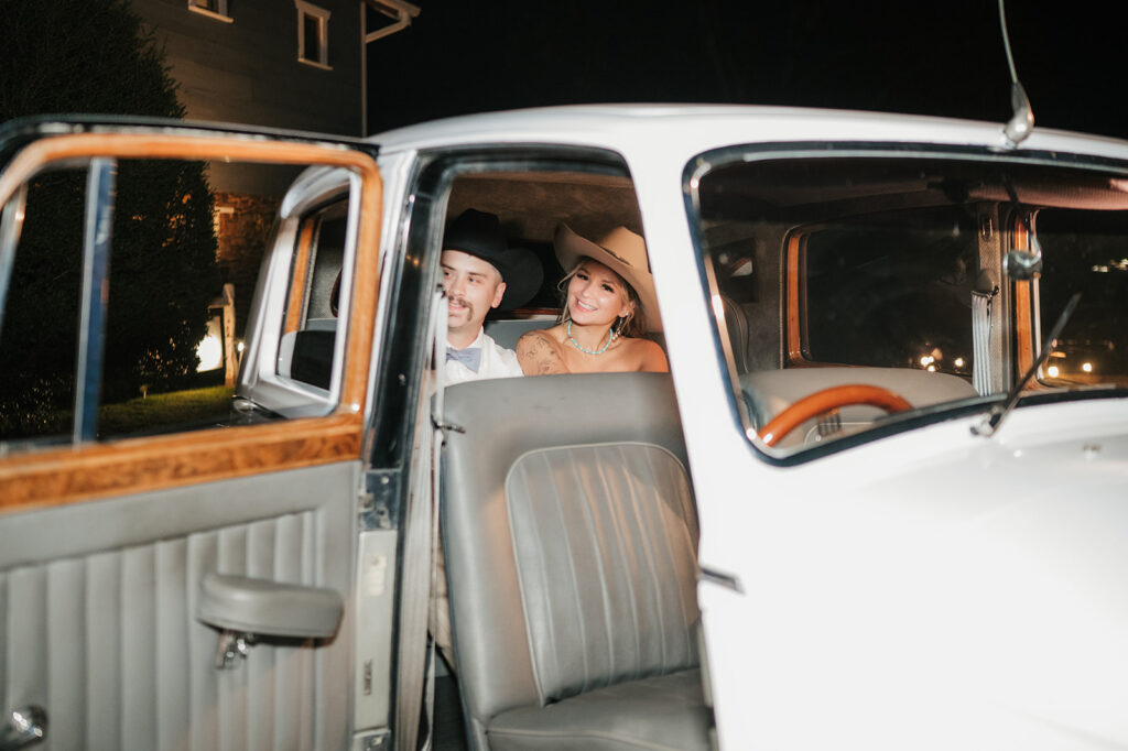 Two people wearing hats sit in the back seat of a vintage white car at night, with the car door open.