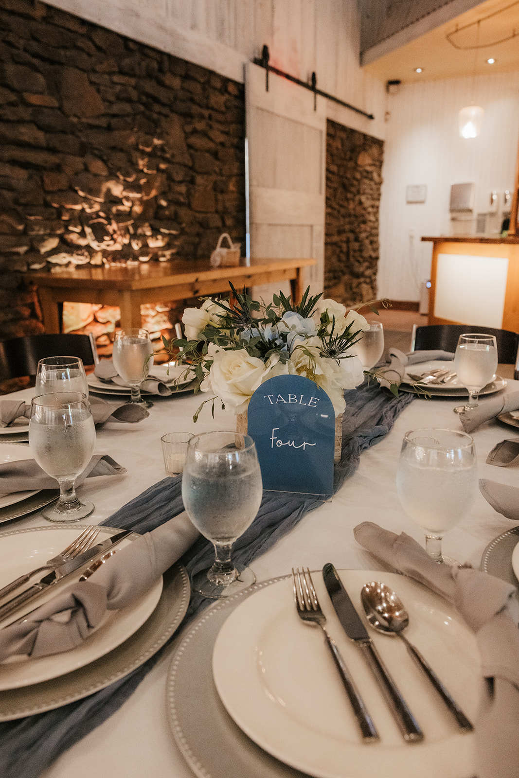 A round table set for a formal event with white plates, silverware, water glasses, cloth napkins, and a flower centerpiece labeled “Table Four.”.