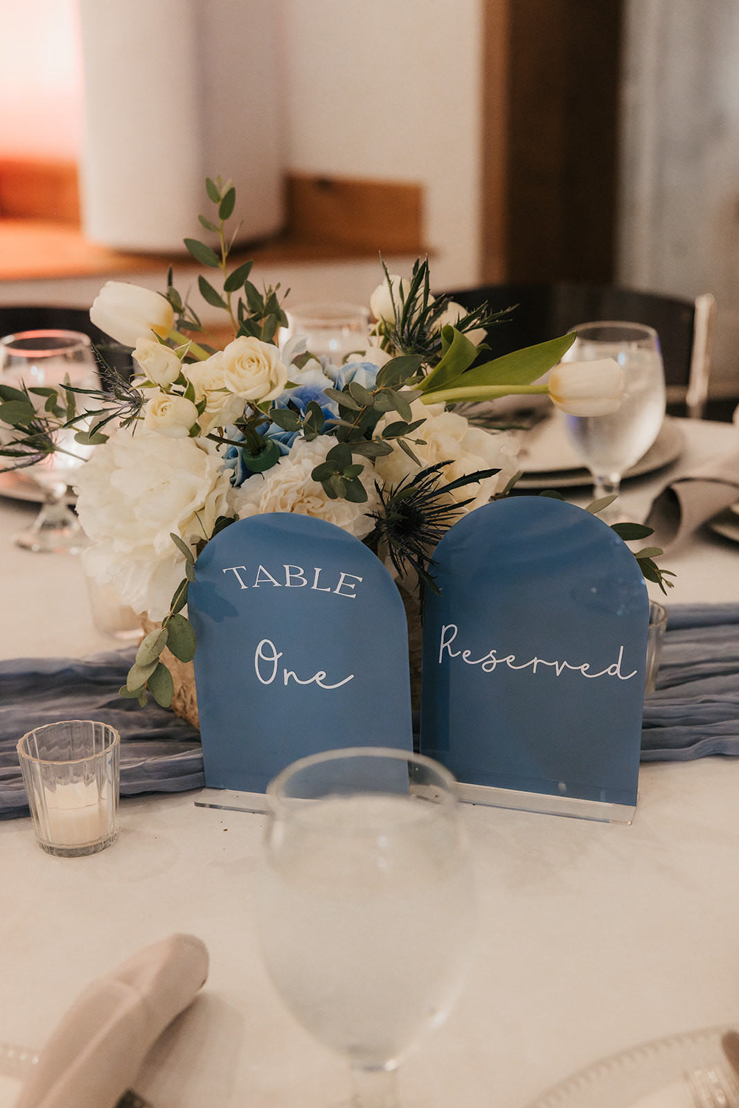 A round table set for a formal event with white plates, silverware, water glasses, cloth napkins, and a flower centerpiece labeled “Table Four.”.