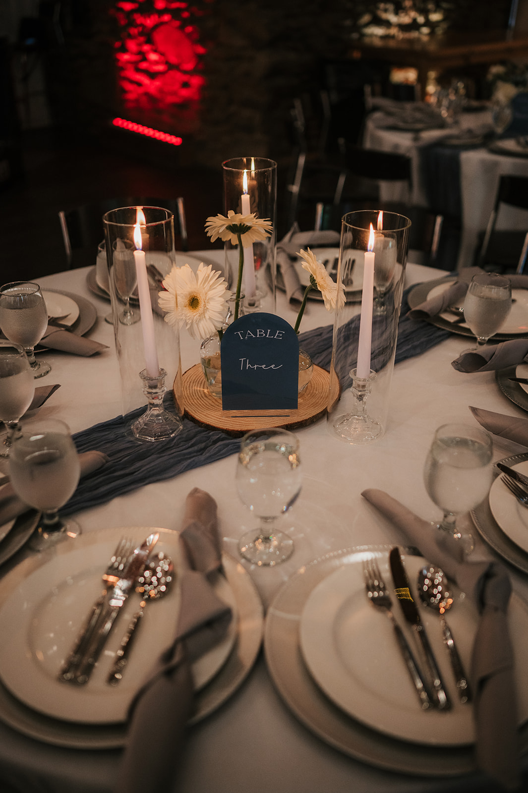 A round table set for a formal event with white plates, silverware, water glasses, cloth napkins, and a flower centerpiece labeled “Table Four.”.